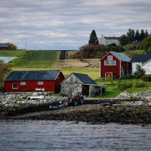Kystgård med røde låver, traktor og jorder ved sjøen i norsk landskap